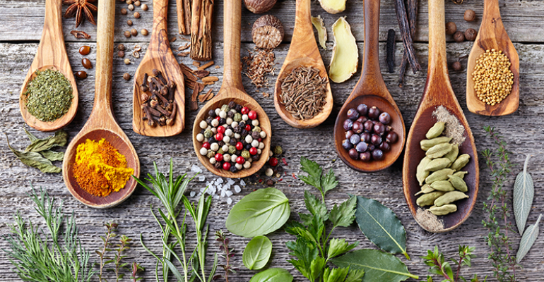 Colheres de madeira posicionadas sobre uma mesa também de madeira, com diversos tipos de temperos para representar as tendências de sabores para 2023.