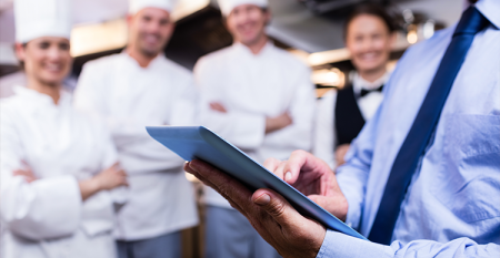 Homem branco vestindo camisa social e gravata azuis, segurando um tablet com a mão esquerda para representar as tecnologias no food service. No fundo da imagem, estão três cozinheiros vestindo uniformes brancos.